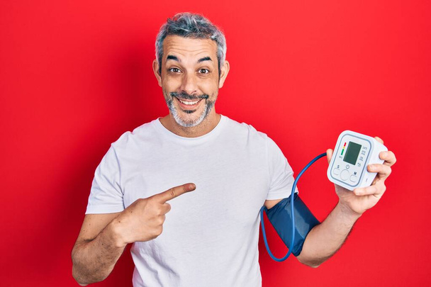 Knappe man van middelbare leeftijd met grijs haar met behulp van bloeddrukmeter glimlachend vrolijk wijzend met hand en vinger  - Foto, afbeelding