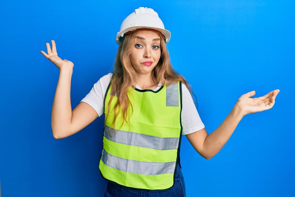 Beautiful blonde caucasian woman wearing architect hardhat clueless and confused with open arms, no idea and doubtful face.  - Photo, image