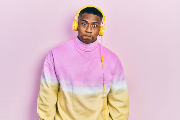 Jonge zwarte man die naar muziek luistert met een koptelefoon die wangen puft met een grappig gezicht. mond opgeblazen met lucht, gekke uitdrukking.  - Foto, afbeelding