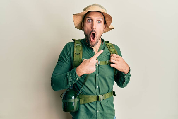 Handsome man with beard wearing explorer hat and backpack surprised pointing with finger to the side, open mouth amazed expression.  - Фото, изображение
