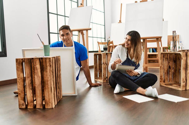 Два студента улыбаются счастливой картиной, сидя на полу в художественной школе. - Фото, изображение