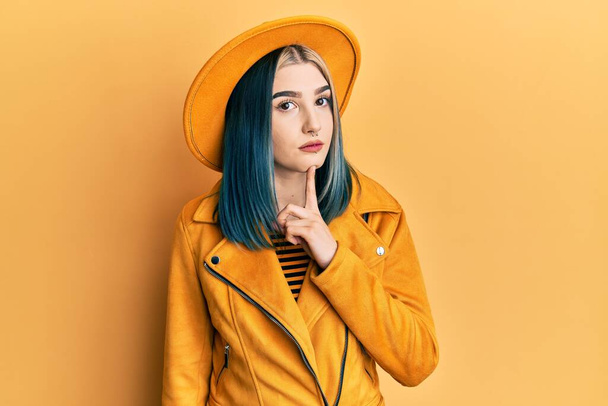 Νεαρό σύγχρονο κορίτσι φορώντας κίτρινο καπέλο και δερμάτινο μπουφάν σκέψης επικεντρώθηκε για την αμφιβολία με το δάχτυλο στο πηγούνι και αναζητούν αναρωτιούνται  - Φωτογραφία, εικόνα