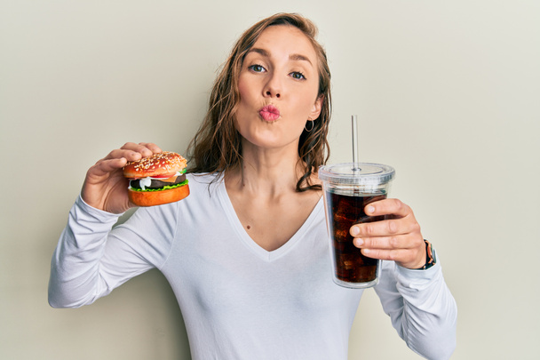 Young blonde woman eating a tasty classic burger with fries and soda looking at the camera blowing a kiss being lovely and sexy. love expression.  - Zdjęcie, obraz