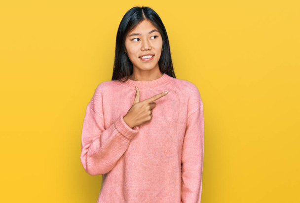 Красивая молодая азиатка в обычном зимнем свитере с улыбкой на лице, указывающей рукой и пальцем в сторону, со счастливым и естественным выражением лица  - Фото, изображение