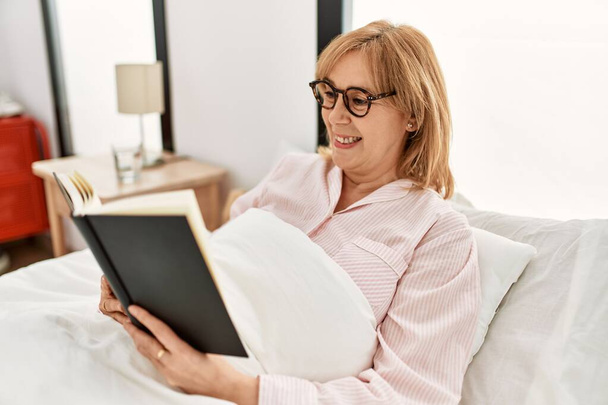 Middle age blonde woman reading book lying on the bed at home. - Foto, Imagen