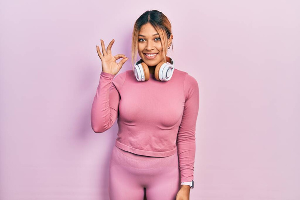 Beautiful hispanic woman wearing gym clothes and using headphones smiling positive doing ok sign with hand and fingers. successful expression.  - Photo, Image