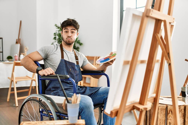 junger hispanischer Mann, der im Rollstuhl im Kunststudio sitzt und Wangen mit lustigem Gesicht aufbläst. Mund mit Luft aufgeblasen, verrückter Ausdruck.  - Foto, Bild