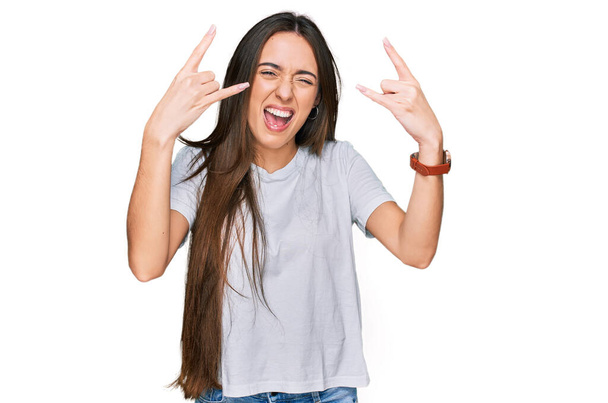 Chica hispana joven con camiseta blanca casual gritando con expresión loca haciendo símbolo de roca con las manos arriba. Estrella de la música. concepto de música pesada.  - Foto, imagen