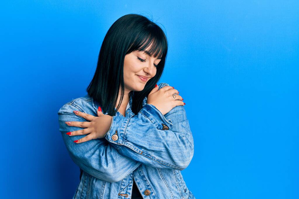 Jeune femme hispanique portant une veste décontractée en denim se serrant dans ses bras heureux et positifs, souriant confiant. amour de soi et soins de soi  - Photo, image