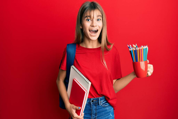 Подростковая кавказка с тетрадками и цветными карандашами празднует безумие и удивляется успеху с открытыми глазами, крича взволнованно.  - Фото, изображение