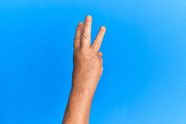 Mano del hombre hispano mayor sobre fondo azul aislado contando el número 3 mostrando tres dedos  - Foto, imagen