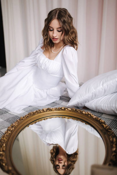 Modelo feminina elegante sentar-se perto do espelho com seu reflexo. Linda e sexy noiva em um vestido curto na cama estirpe seu cabelo - Foto, Imagem
