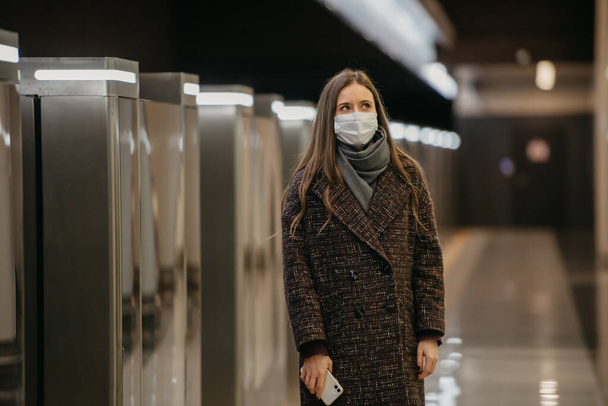 La señora con una máscara facial médica para evitar la propagación del coronavirus está esperando un tren y sosteniendo un teléfono celular en la plataforma del metro. Una chica con una máscara quirúrgica mantiene una distancia social en el metro - Foto, imagen