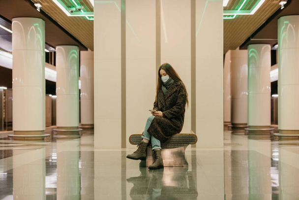 Uma mulher solitária em uma máscara facial médica para evitar a propagação do coronavírus está sentada usando um smartphone na plataforma do metrô. Uma menina em uma máscara cirúrgica está mantendo a distância social no metrô. - Foto, Imagem