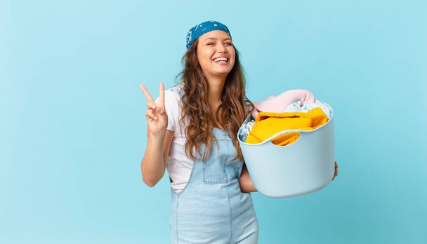 young pretty woman smiling and looking friendly, showing number two and holding a wash clothes basket - Photo, image
