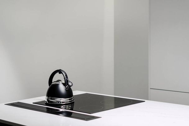 Weiße Arbeitsplatte mit kleinem schwarzen Wasserkocher auf eingebauter elektrischer Induktionsherdplatte und weißer Kubatur dahinter in sauberer, moderner, leerer Küche mit niemandem - Foto, Bild