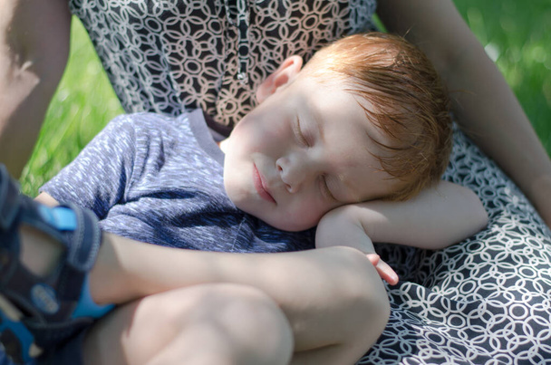 L'enfant dort en plein air sur les genoux de la mère. Le sommeil des enfants dans la nature. Un garçon roux de quatre ans dort dans les bras de sa mère - Photo, image