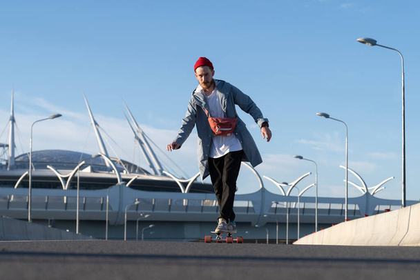 Lässiger Skater-Mann auf dem Longboard im Stadtraum. Hipster-Mann trägt trendige Straßenmode - Foto, Bild