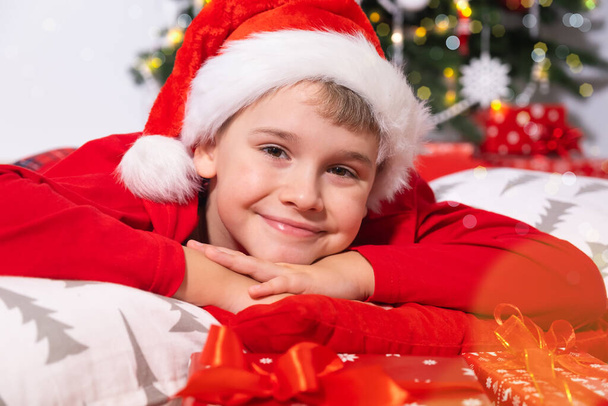 Vacaciones de Navidad. Retrato del niño sonriente en Santa Sombrero Rojo mirando a la cámara, yace rodeado de regalos de Navidad. - Foto, Imagen