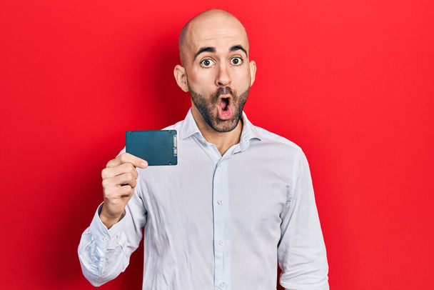 Young bald man holding ssd memory scared and amazed with open mouth for surprise, disbelief face  - Photo, Image