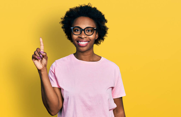 Afrikaans amerikaanse vrouw met afro haar dragen casual kleding en bril tonen en wijzen met vinger nummer een terwijl glimlachen zelfverzekerd en gelukkig.  - Foto, afbeelding