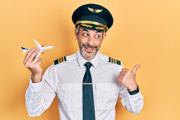 Красивый мужчина средних лет с седыми волосами в форме пилота самолета держит игрушечный самолет указывая большой палец вверх в сторону улыбаясь счастливым с открытым ртом  - Фото, изображение