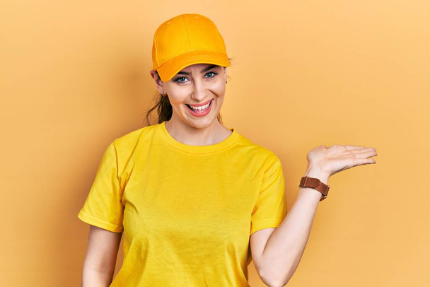 Jonge Latijns-Amerikaanse vrouw draagt levering uniform en pet glimlachend vrolijke presenteren en wijzen met palm van de hand kijken naar de camera.  - Foto, afbeelding