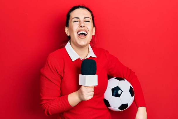 Young hispanic woman holding reporter microphone and soccer ball smiling and laughing hard out loud because funny crazy joke.  - Photo, Image