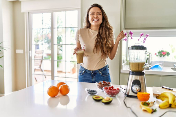 Beautiful young brunette woman drinking glass of smoothie at the kitchen screaming proud, celebrating victory and success very excited with raised arms  - Photo, Image