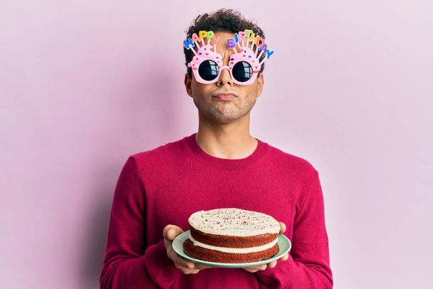 Handsome hispanic man wearing funny happy birthday glasses holding cake skeptic and nervous, frowning upset because of problem. negative person.  - Photo, Image