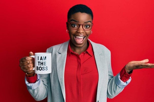 Jovem afro-americana bebendo de eu sou o chefe xícara de café celebrando realização com sorriso feliz e expressão vencedora com mão levantada  - Foto, Imagem