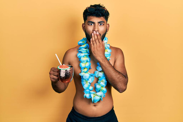 Arab man with beard wearing swimsuit and hawaiian lei drinking cocktail covering mouth with hand, shocked and afraid for mistake. surprised expression  - Foto, Bild