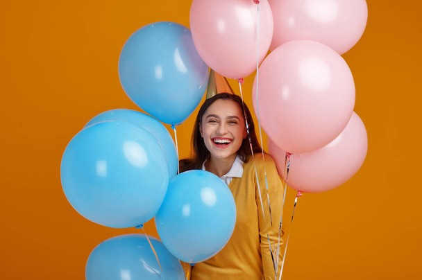 Funny woman in cap, yellow background. Pretty female person got a surprise, event or birthday celebration, balloons - Фото, изображение
