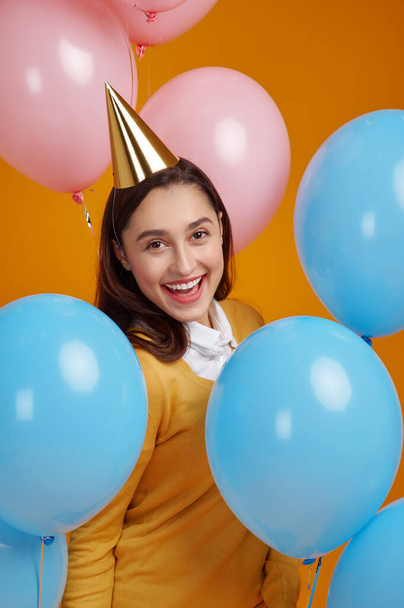 Funny woman in cap, yellow background. Pretty female person got a surprise, event or birthday celebration, balloons decoration - Photo, image
