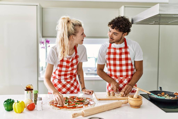 Νεαρό ζευγάρι χαμογελά ευτυχισμένη μαγείρεμα ιταλική πίτσα στην κουζίνα. - Φωτογραφία, εικόνα