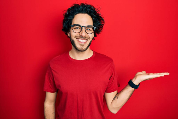 Knappe latino man met casual t-shirt en bril lachend vrolijke presentatie en wijzend met handpalm kijkend naar de camera.  - Foto, afbeelding