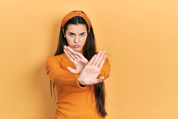 Joven adolescente morena con expresión de rechazo casual suéter amarillo cruzando brazos y palmas haciendo signo negativo, cara enojada  - Foto, Imagen