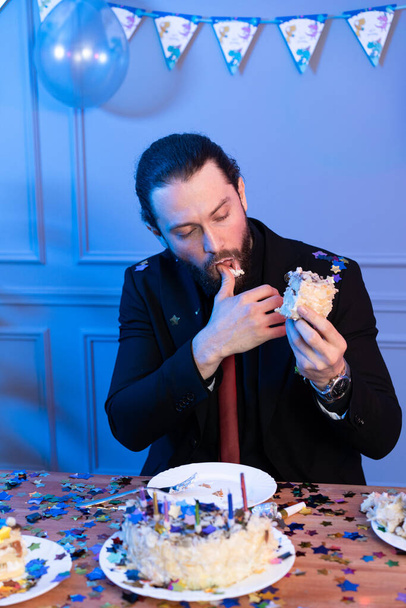 The man is sitting at the table holding a piece of cake and looking at it. Atypical birthday celebration. - Foto, Imagem