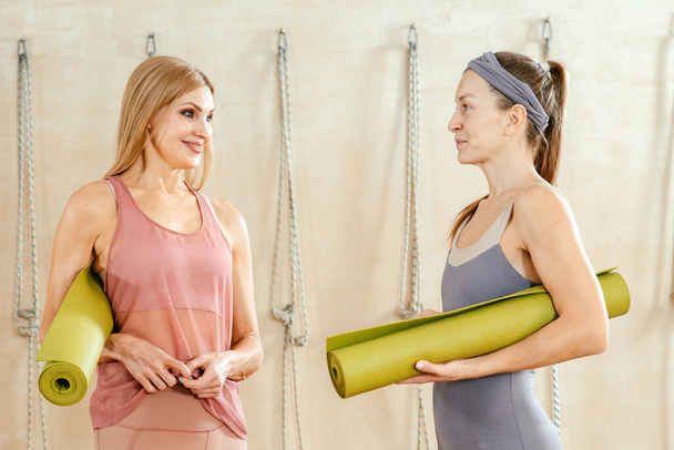 Two women wearing activewear standing in gym holding yoga mat ready to start fitness or yoga training. Concept of sports lifestyle. Yoga concept - Photo, image