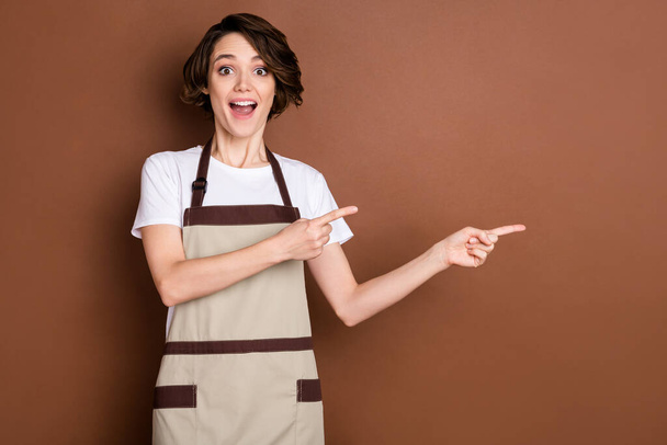Portrait of attractive amazed cheerful girl barista coffee shop demonstrating copy space advert isolated over brown color background - Foto, Imagen