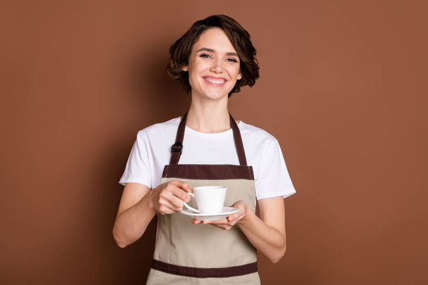 Portrait of attractive cheerful girl holding in hands bringing espresso serving tables isolated over brown color background - Fotó, kép