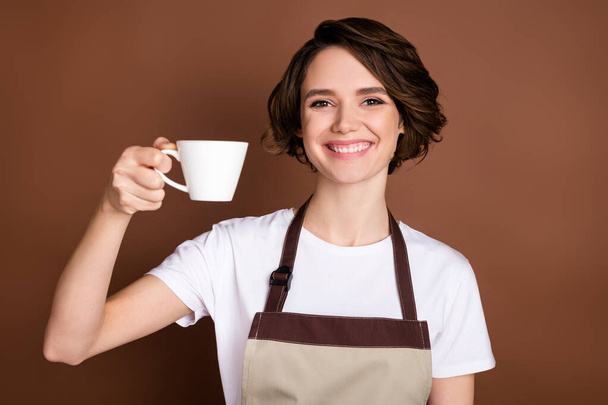 Foto de la mano dama positiva sosteniendo taza de café sonrisa dentada mirada cámara aislada sobre fondo de color marrón - Foto, imagen