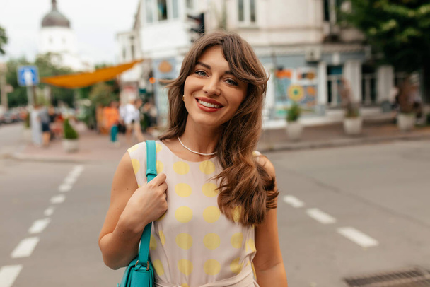 Joyeux adorable fille avec les cheveux ondulés en tenue d'été posant à la caméra sur le fond de la rue de la ville dans la chaude journée d'été. Photo de haute qualité - Photo, image