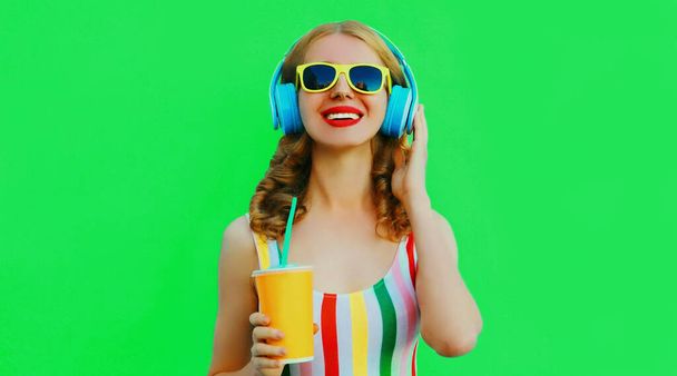 Portrait of happy smiling young woman listening to music in headphones with fresh cup of juice on green background - Фото, изображение