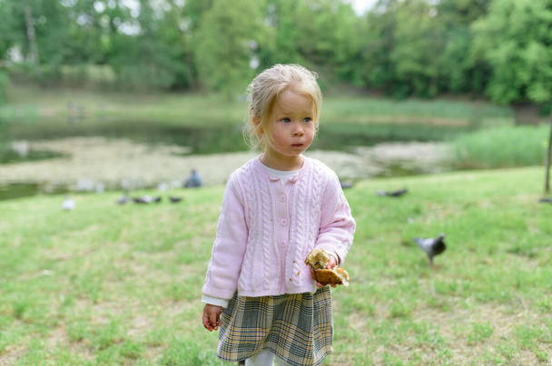 Uma menina de 2 anos de idade com lábios amuados, segura um pão na mão. - Foto, Imagem