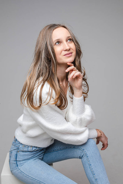 Портрет молодої мрійливої довгошерстої білої дівчини в білому светрі і блакитних джинсах на сірому студійному фоні
 - Фото, зображення