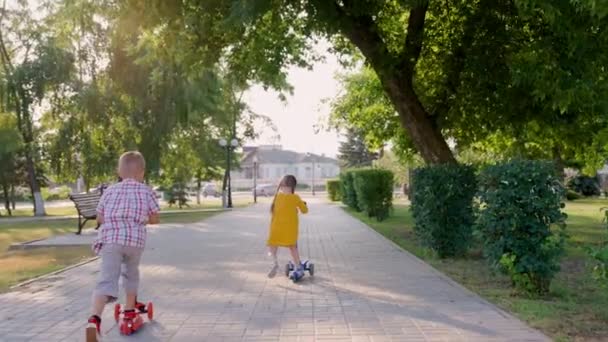Chica y niño montar scooter juntos al aire libre. Feliz lindo niños jugando en el parque en la calle, aprendiendo a equilibrar en scooter. El niño y la niña juegan despreocupados en el parque infantil. Familia fin de semana verano - Metraje, vídeo