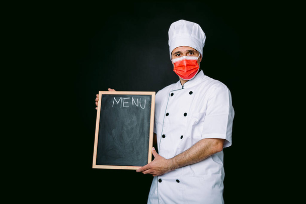 Chef cook wearing cooking jacket and hat with red mask, holding a blackboard with menu, on black background - Photo, Image