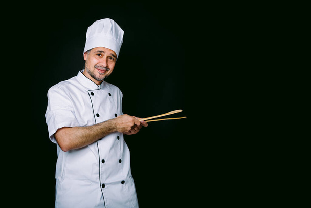 Chef cocinero con chaqueta y sombrero de cocina, sosteniendo cuchara de madera y tenedor, apuntando a su izquierda, sobre fondo negro - Foto, Imagen
