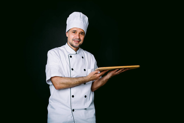 Cuoco cuoco indossa giacca da cucina e cappello, in possesso di un vassoio di legno, su sfondo nero - Foto, immagini